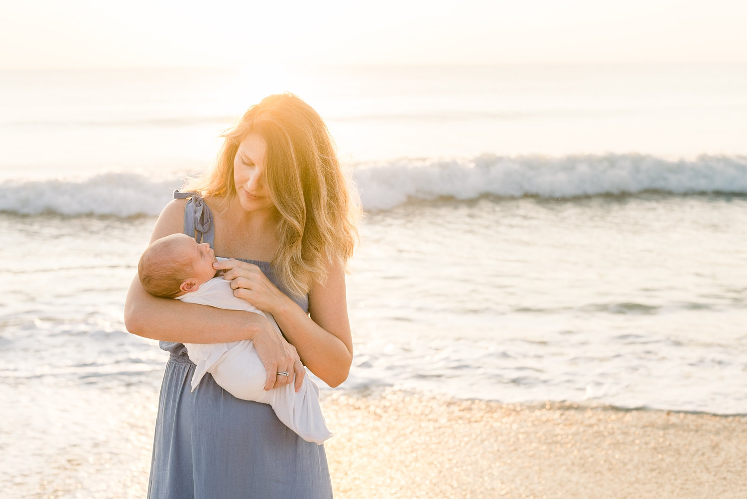 mom wearing a cornflower blue dress, she is holding her newborn baby, the sun is rising in the background