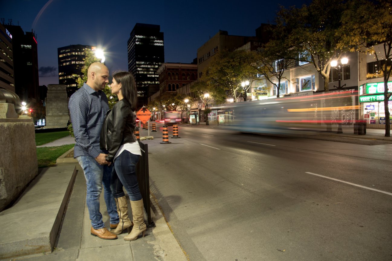 Couple embracing on a city sidewalk at night; blurred traffic and illuminated buildings in the background. Hamilton, On