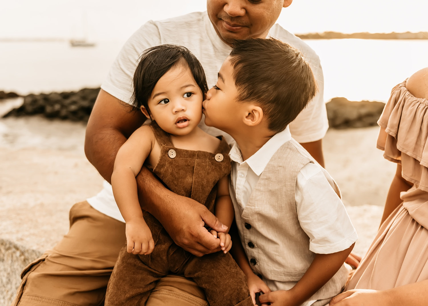 father holding his 2 young boys, one brother is kissing the other brother's cheek, mom's dress is peeking from the right side of the image