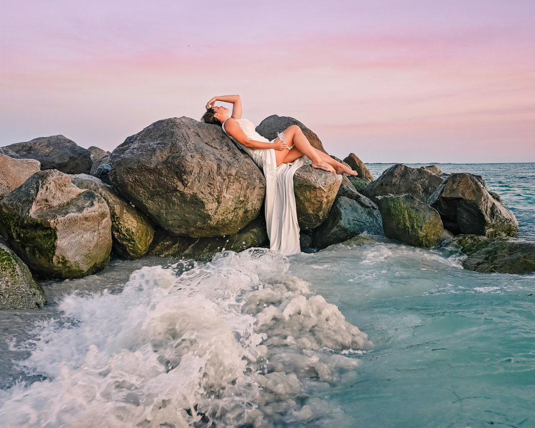 A woman in a flowing white dress poses on large rocks at the beach with gentle waves and a pastel sky.