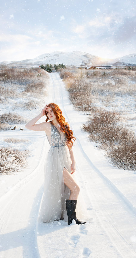 Young woman in a glittering gown poses on a snowy path, surrounded by winter scenery. Snow gently falls around her.
