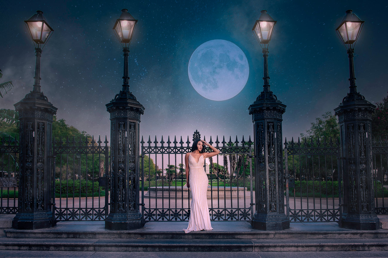 A woman in a white dress stands by an ornate gate under a full moon at night, with street lamps illuminating the scene.