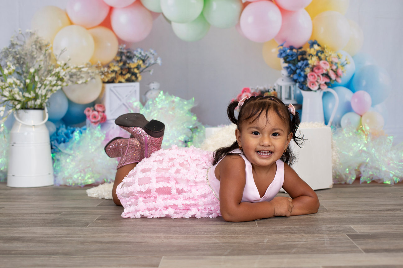 Smiling toddler girl lying on a wooden floor in front of a pastel birthday balloon backdrop.