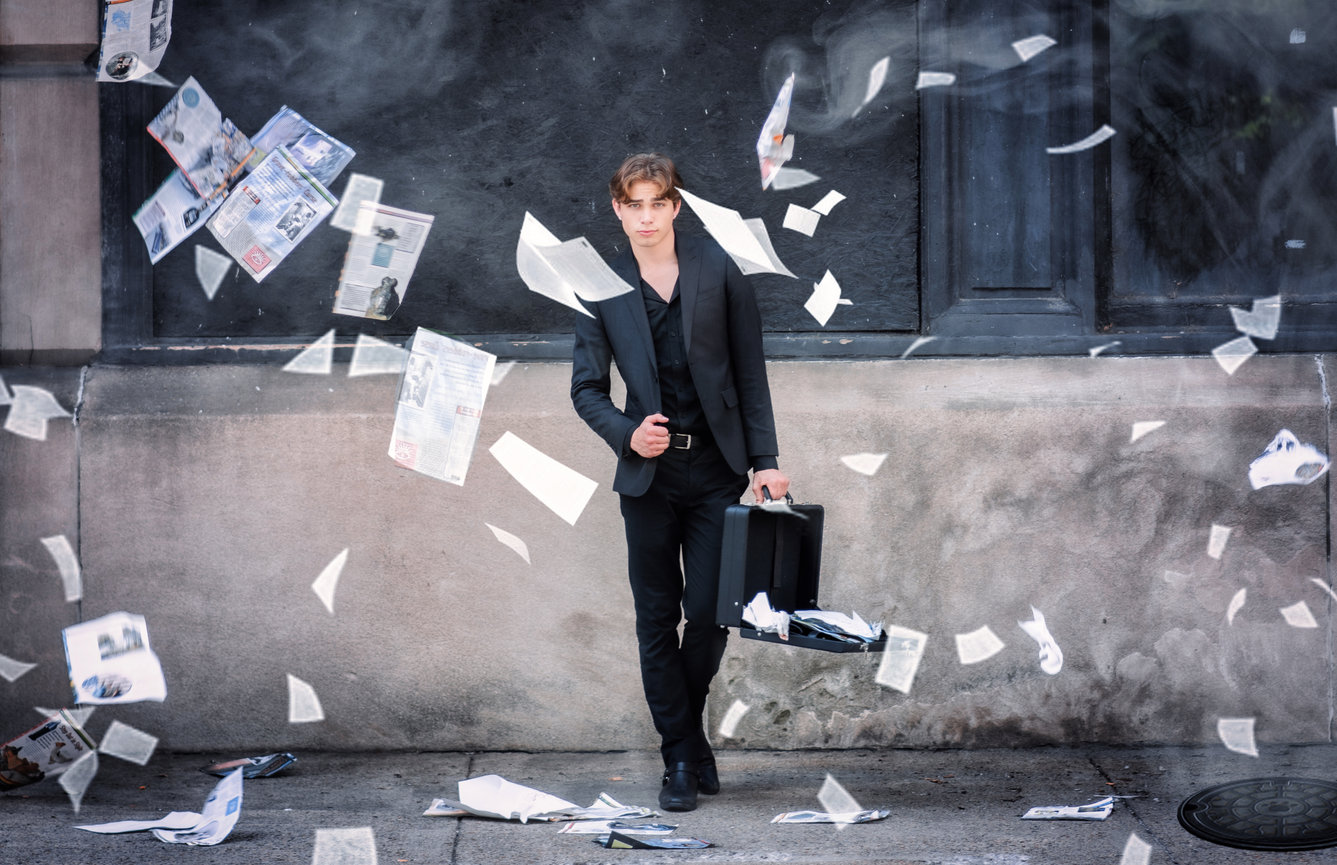 A man in a black suit stands against a wall, holding a briefcase as papers swirl around him.