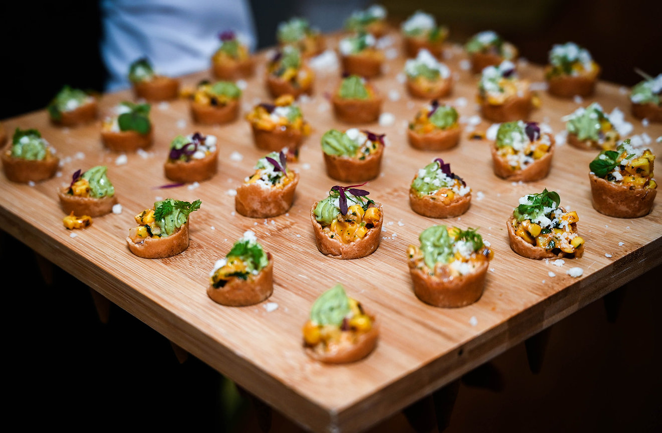 A wooden platter filled with mini pastry cups topped with guacamole, corn, and herbs.