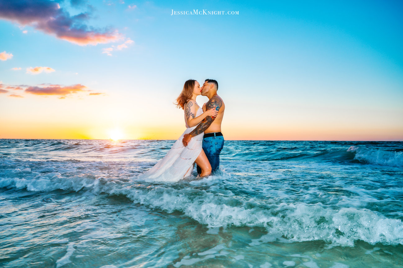 Couple kissing in the ocean at sunset, with waves gently crashing around them.