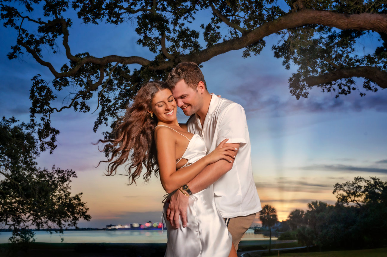 A happy couple embraces outdoors at sunset, surrounded by trees, creating a romantic atmosphere.