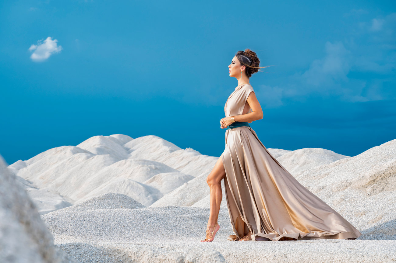 Model in a flowing beige gown poses against a backdrop of white sand dunes under a blue sky.