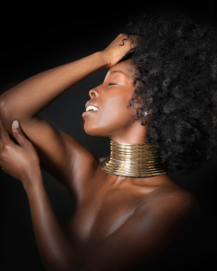 A woman with natural hair poses confidently, wearing a large golden necklace against a dark background.