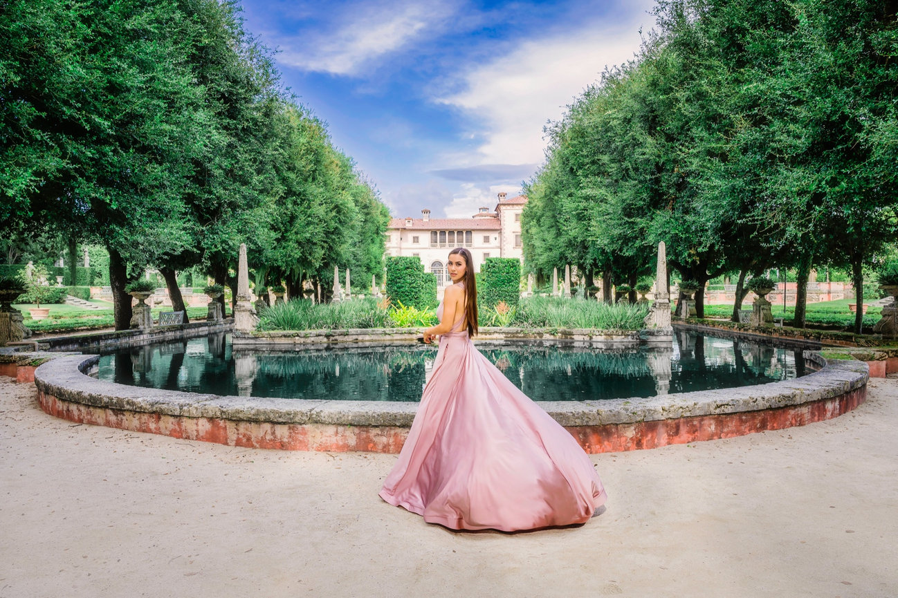 A woman in a flowing pink gown stands by a circular pond in a lush garden, with a grand building in the background.