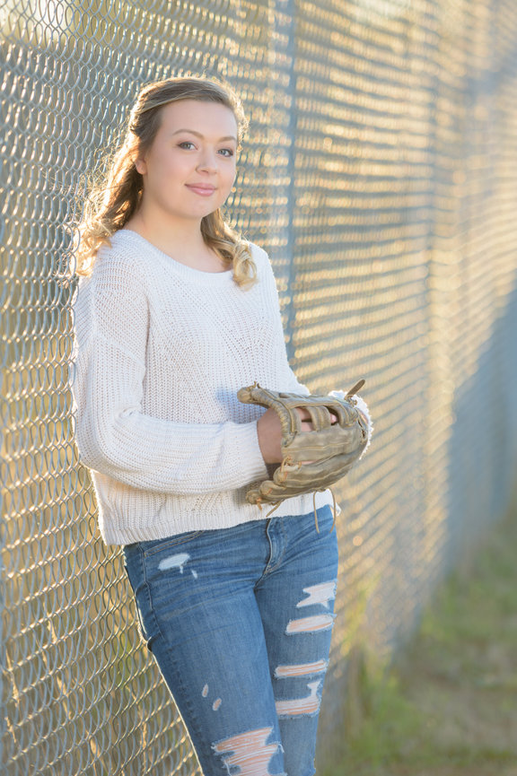 Senior girl leaning against a chainlink fence holding a softball and glove with golden light streaming through the fence lighting her hair