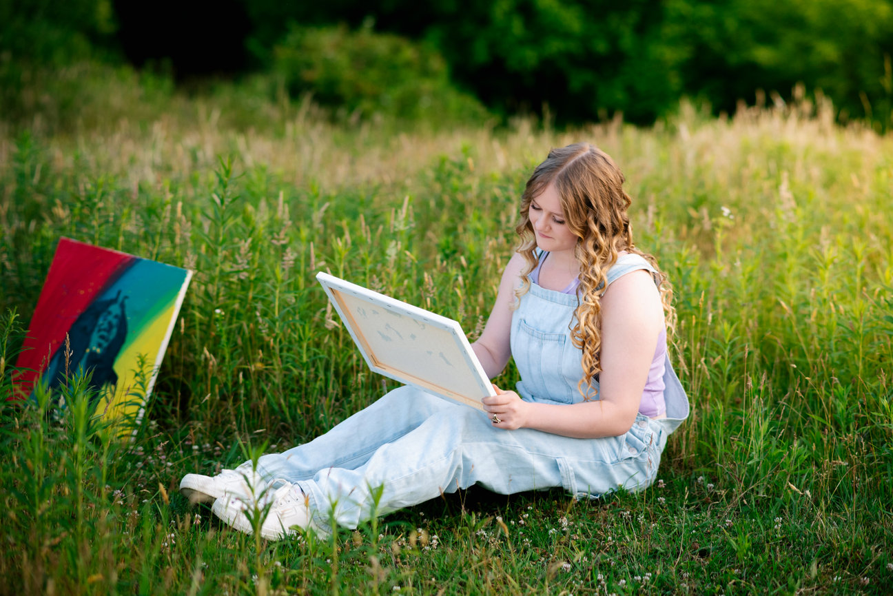 senior girl wearing overalls in a field of high grass working on a painting