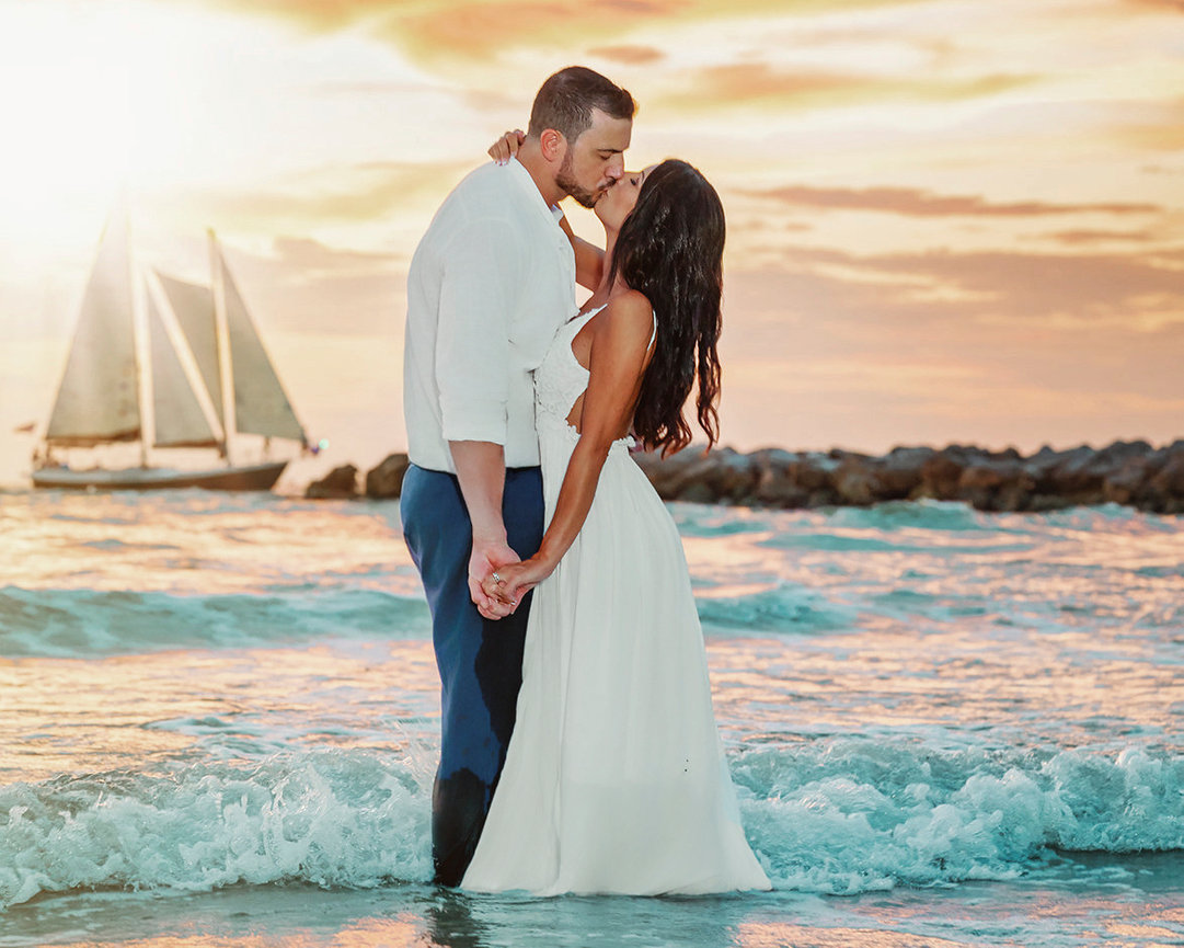 Couple kissing in the ocean at sunset, with sailboats in the background and waves gently splashing.