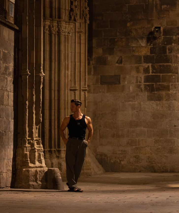 Man in a sleeveless shirt and pants leans against an ornate stone wall in dim lighting. Barcelona night