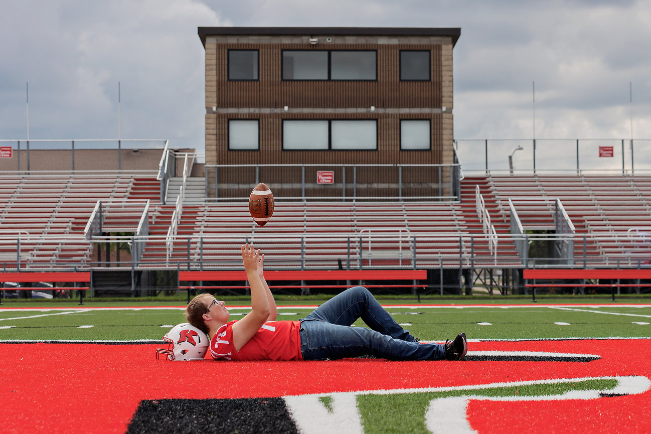 A person lying on a football field, tossing a football, with bleachers in the background.