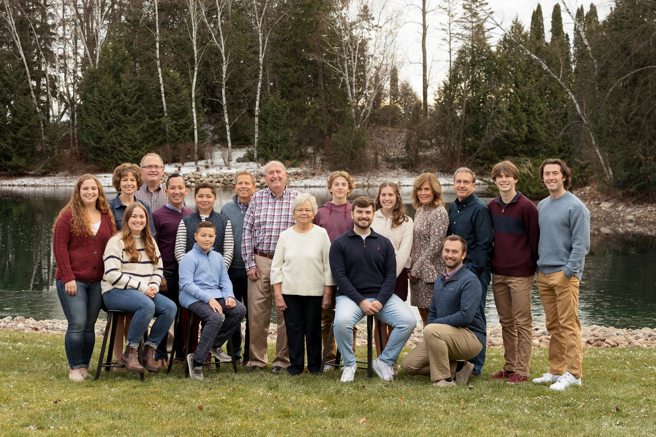 A large family is gathered outdoors near a pond and trees, posing for a group photo on a grassy area.