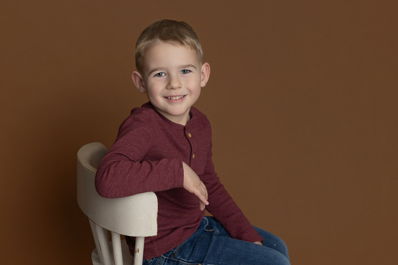 Smiling child in a maroon shirt sits on a white chair against a brown background.