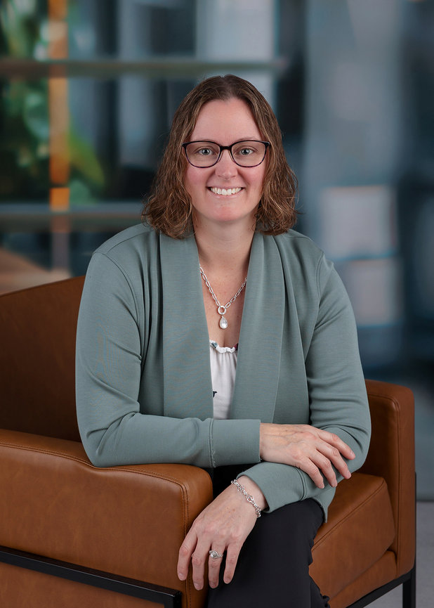 A person with glasses, wearing a green cardigan, sitting on a brown chair, smiling indoors.