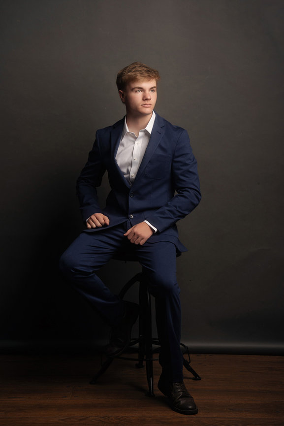 Young man in a blue suit sits on a stool against a dark background, looking to the side.