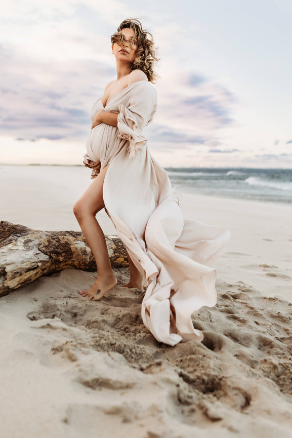 A mother to be in an ivory dress stands on a New Jersey beach during her maternity photo session.