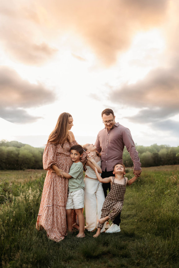 A family with three children standing close together in a field at the Central Park of Morris County. Captured by New Jersey Family Photographer Love of James Photography.
