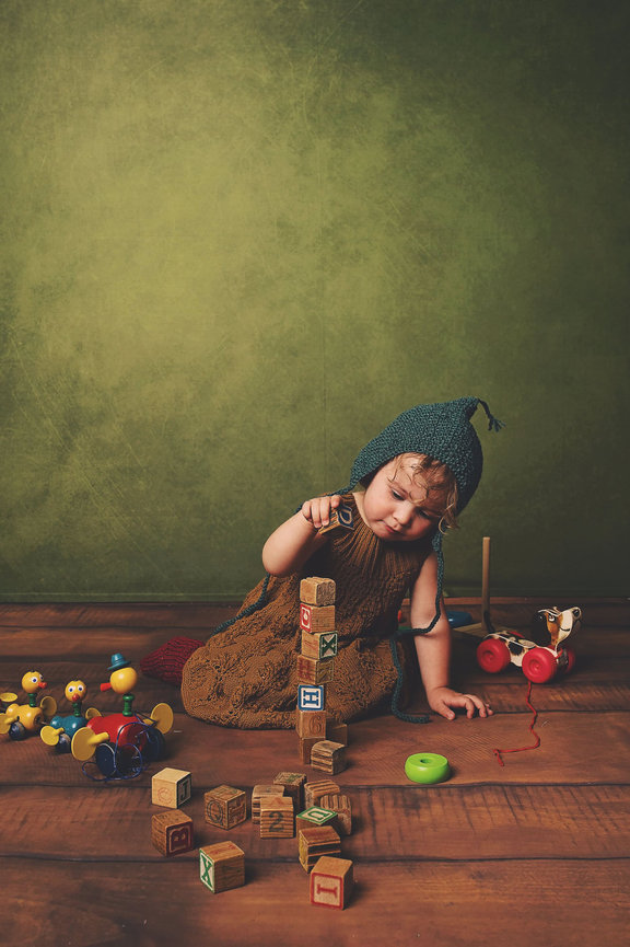 A toddler girl wearing a mustard colored knit dress, red knit socks and a teal knit bonnet stacks blocks while surrounded by vintage toys
