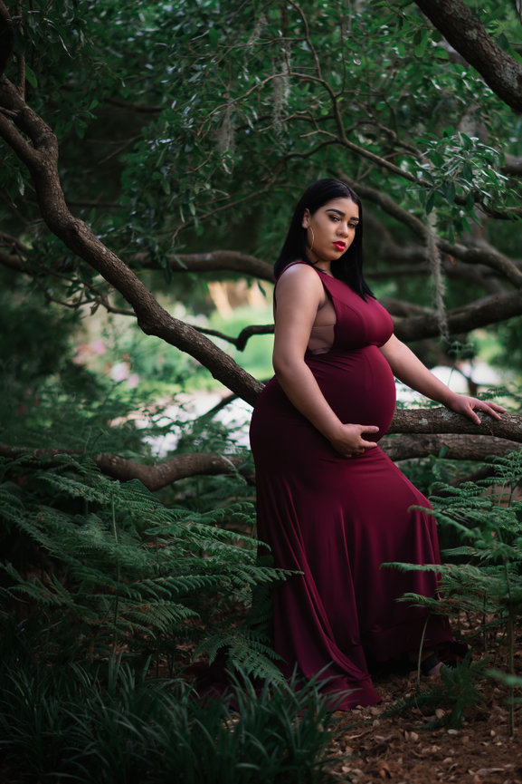 Pregnant woman in a flowing burgundy dress poses in a lush, green forest setting.
