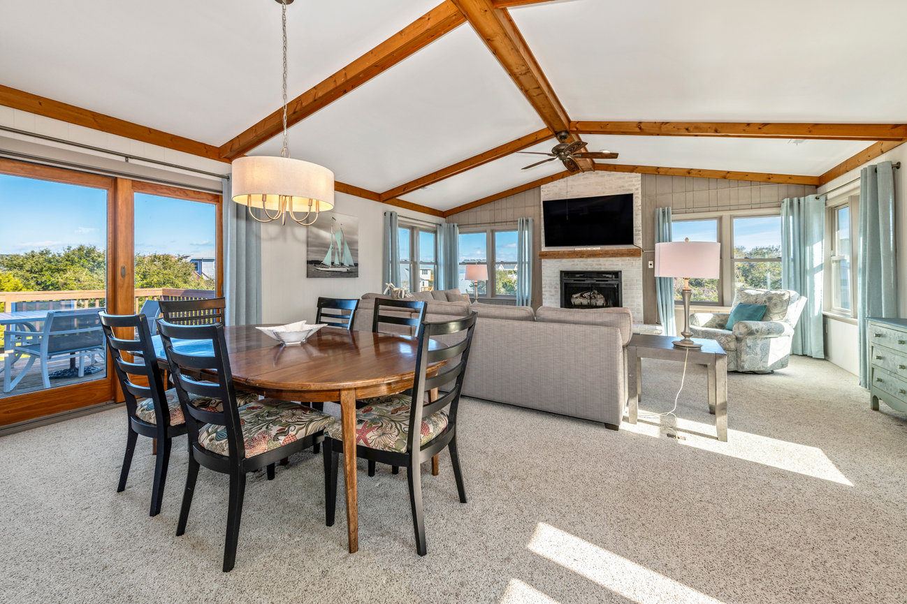 Open-concept living room with wooden beams, dining table, sofa, fireplace, and large windows with a scenic view.