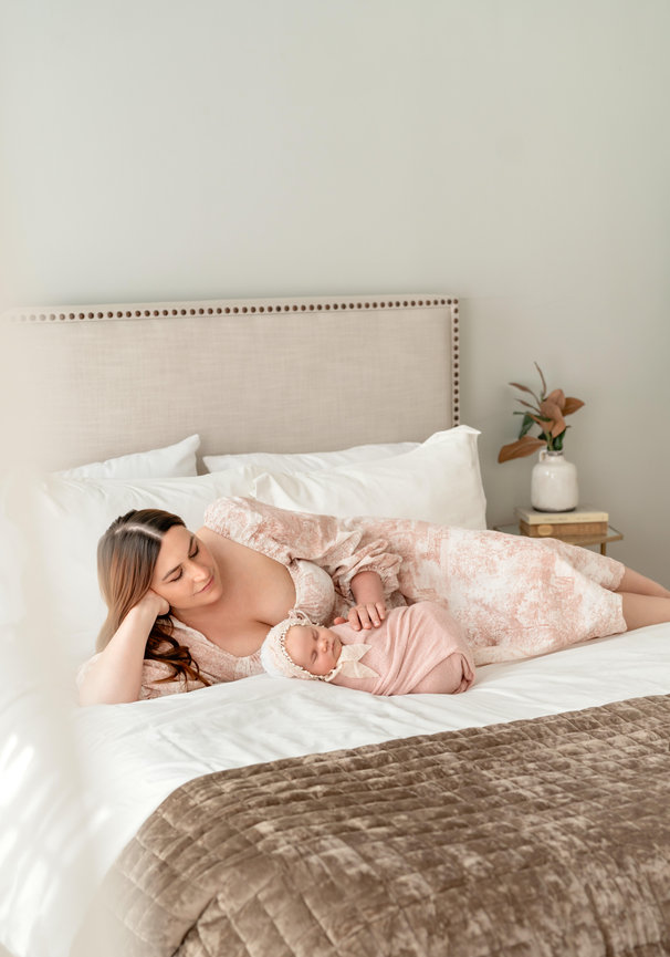 Mother resting with sleeping newborn on a bed in a soft, neutral-toned bedroom
