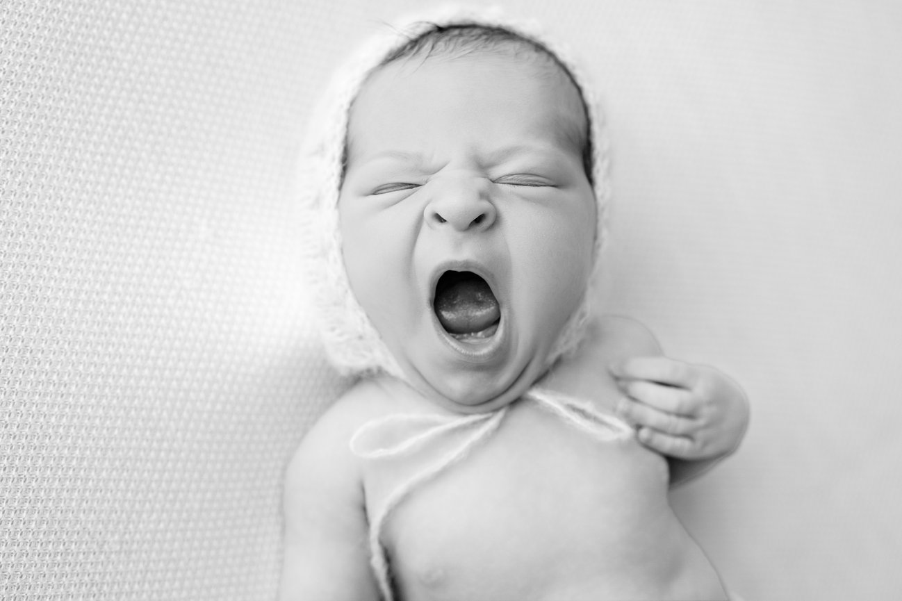 Black and white close-up of a baby yawning, wearing a headband with a bow, lying on a soft blanket — newborn photography detail shot
