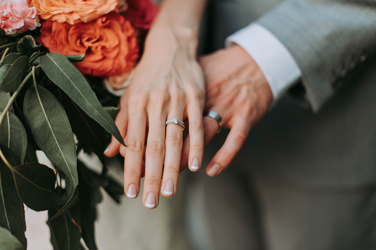 Close-up of wedding rings on intertwined hands beside a bouquet of peach roses.