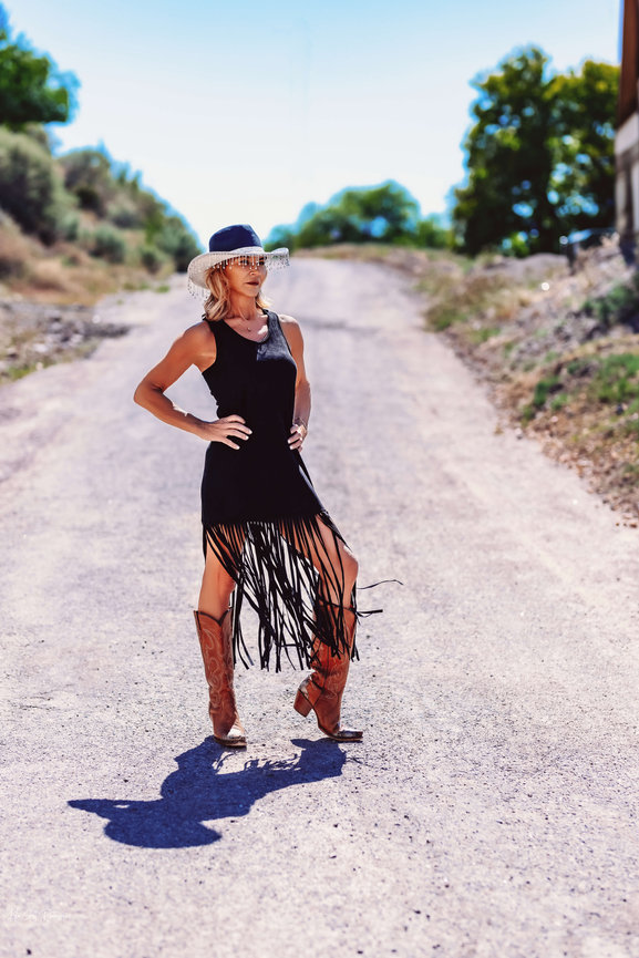 Woman in a black fringe dress and cowboy boots standing on a sunny rural road, wearing a hat.