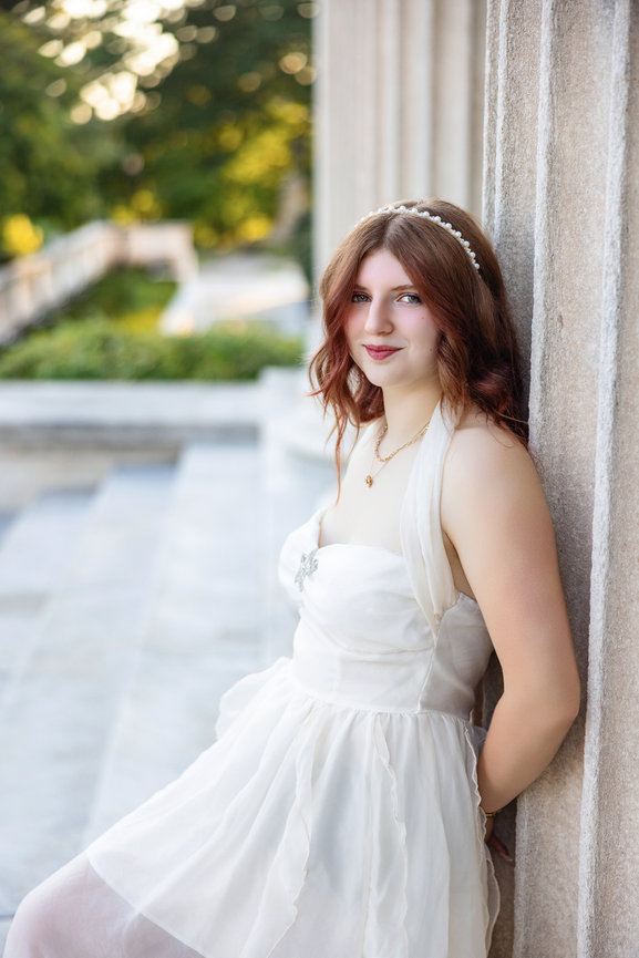High school senior girl in a white dress leaning against a stone column for senior portraits near Cleveland Ohio.