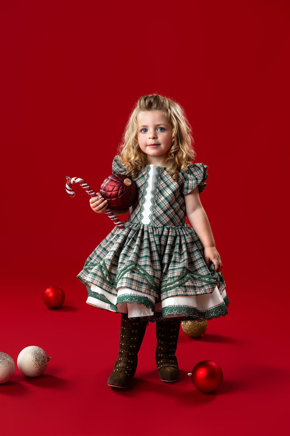 A young girl in a plaid holiday dress holding a large ornament against a classic red Christmas portrait backdrop in Northeast Ohio
