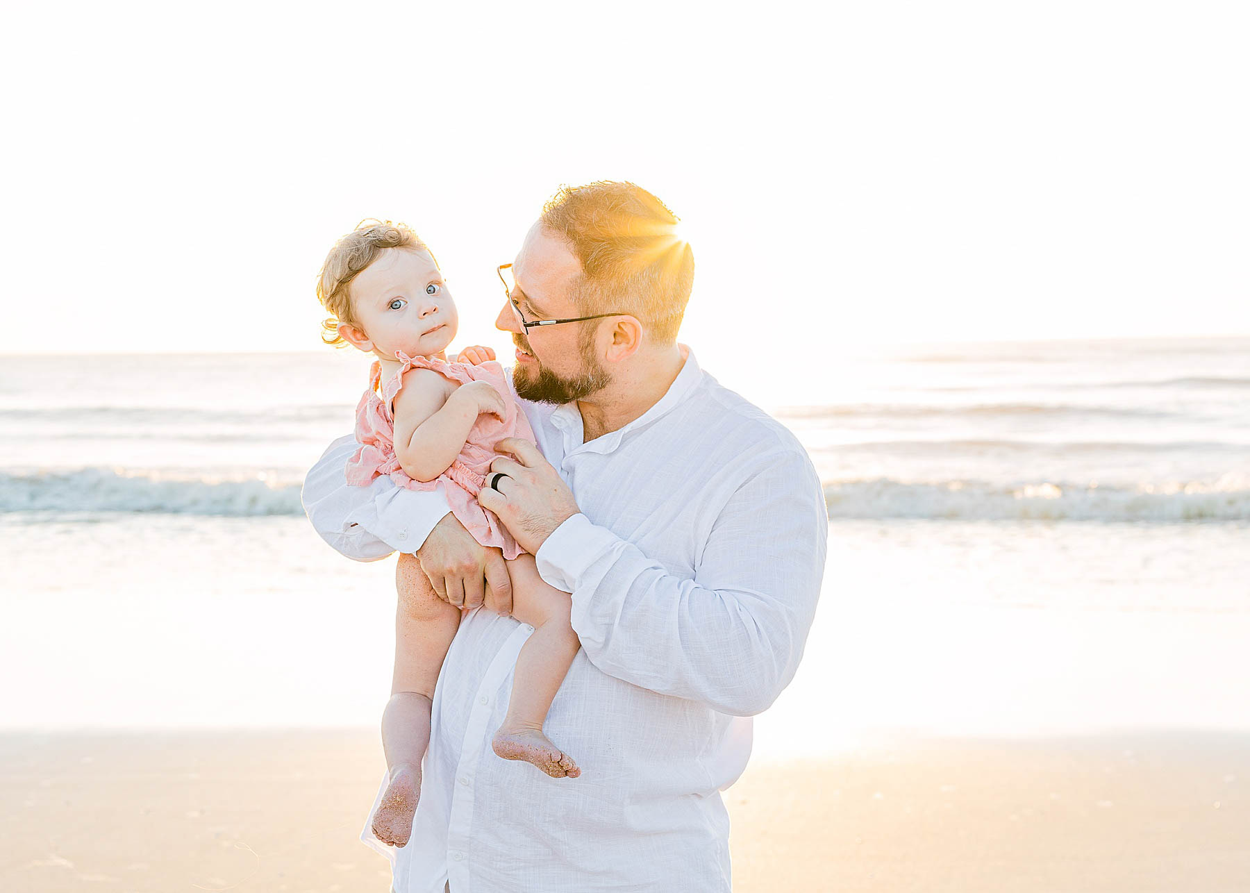 man holding baby wearing a pink dress on the beach at sunrise
