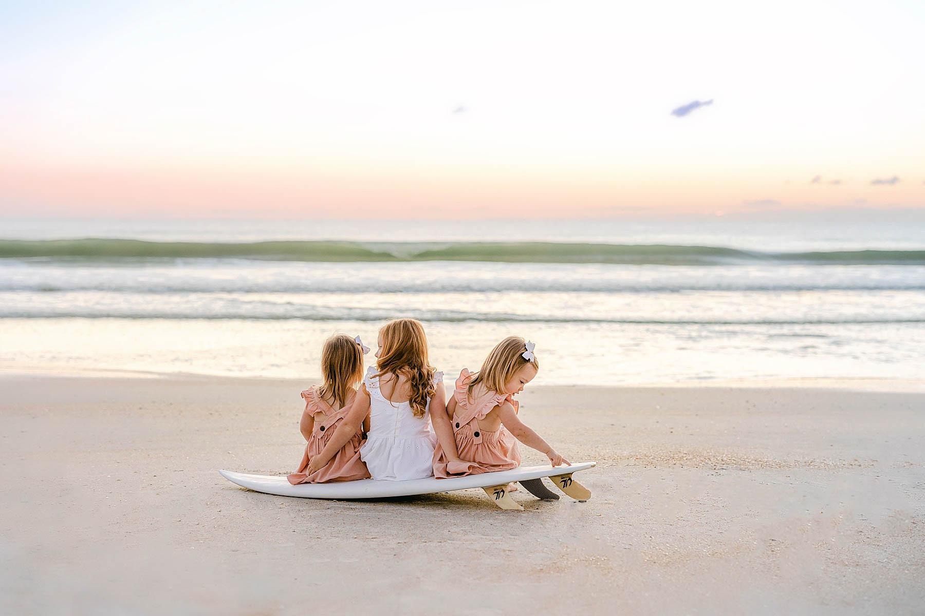 three little girls in dresses sitting on a surfboard at sunrise on the beach