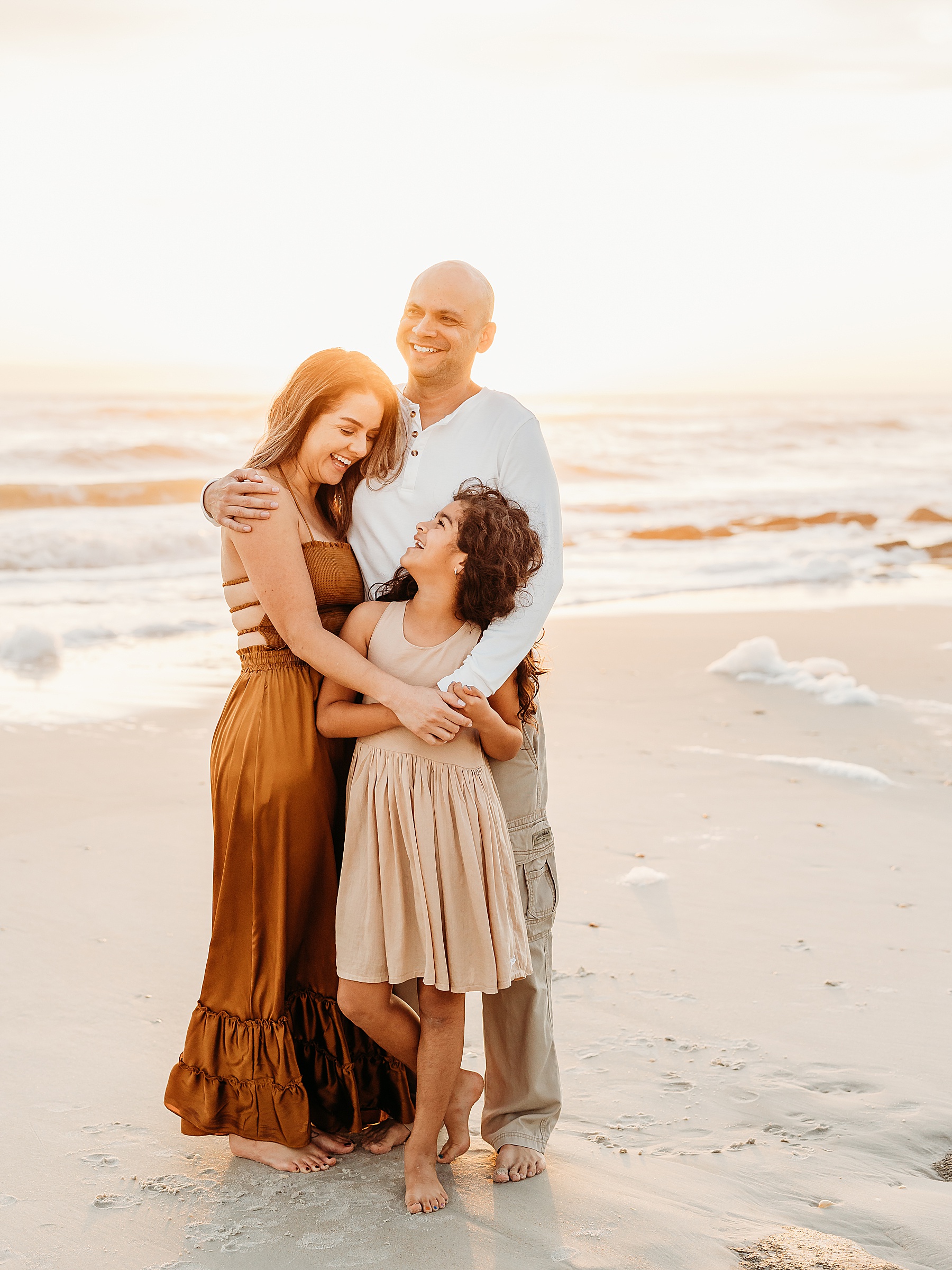 family of three standing near the ocean wearing neutral colors and rust at sunrise