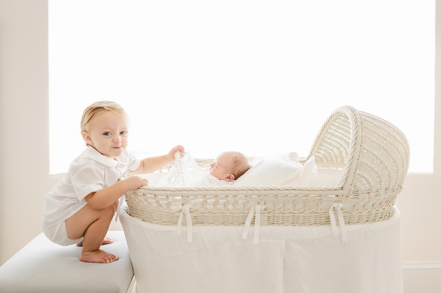 baby boy dressed in white peeking under covers of newborn baby girl in bassinet in white nursery