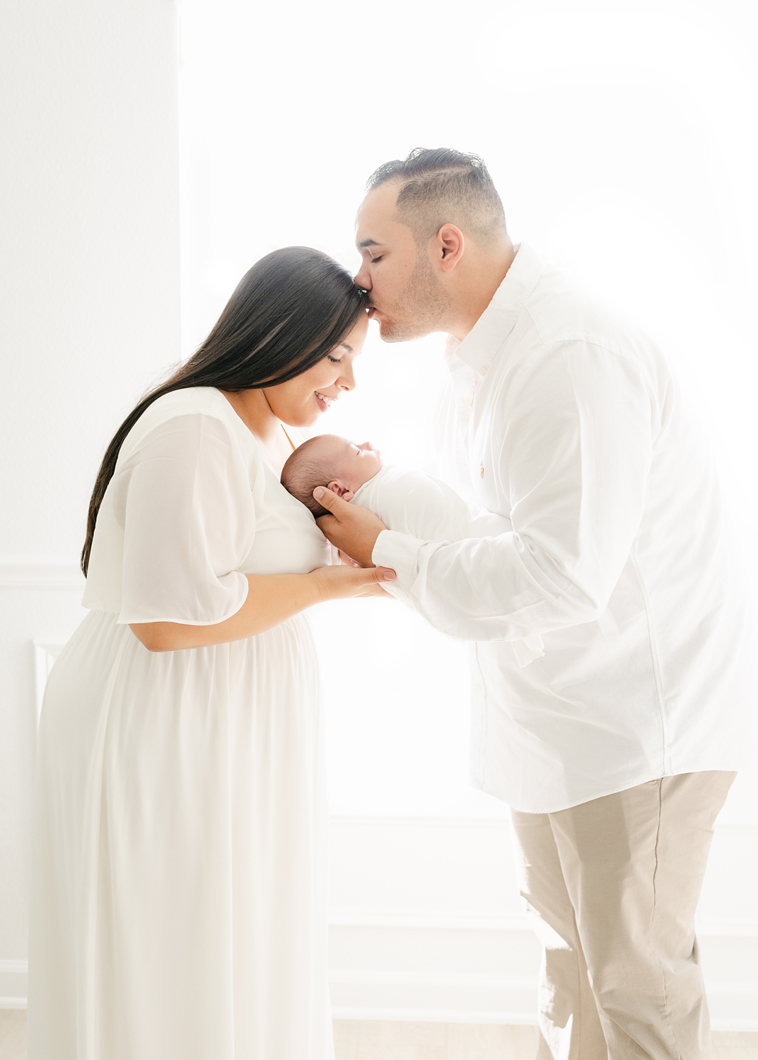 man and woman holding newborn baby boy wrapped in white blanket in all white room
