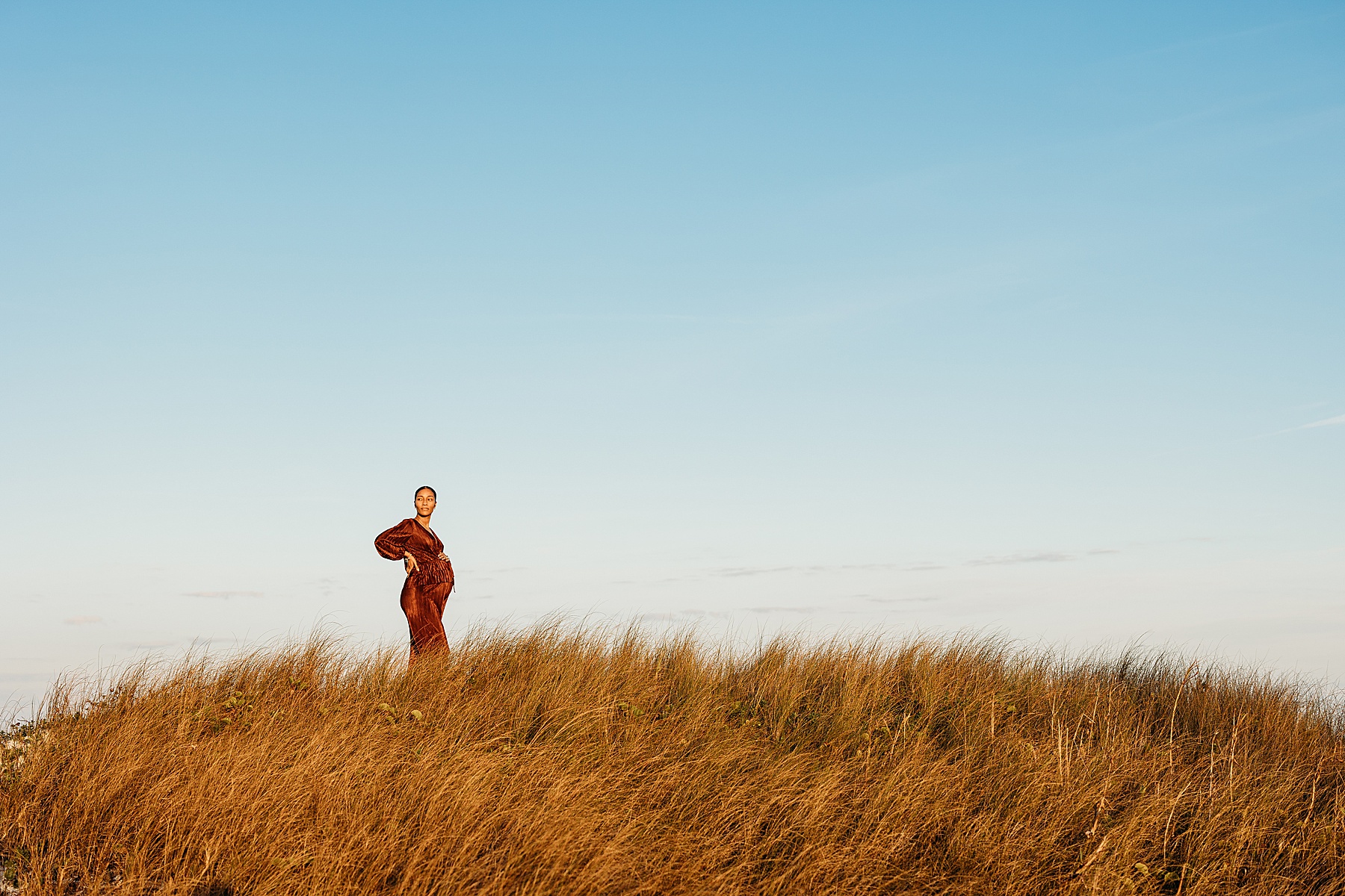 pregnant woman wearing a rust jumpsuit standing on a hill at a golden sunset