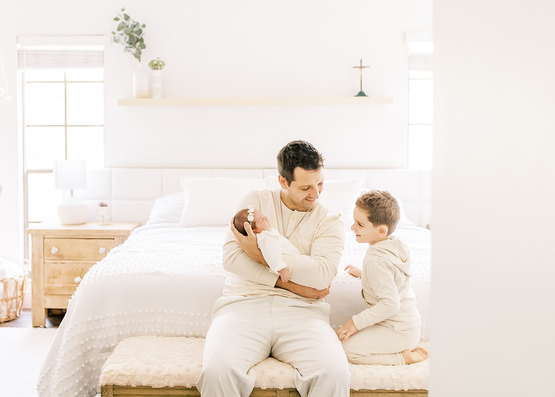 man dressed in cream neutral colors holding baby girl next to little boy dressed in tan in a white room