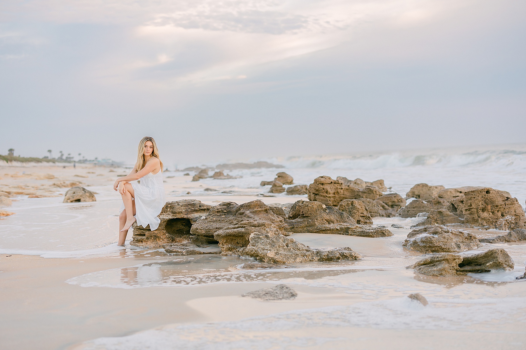 young blond woman in long white maxi dress sitting on rocks at the beach at sunrise