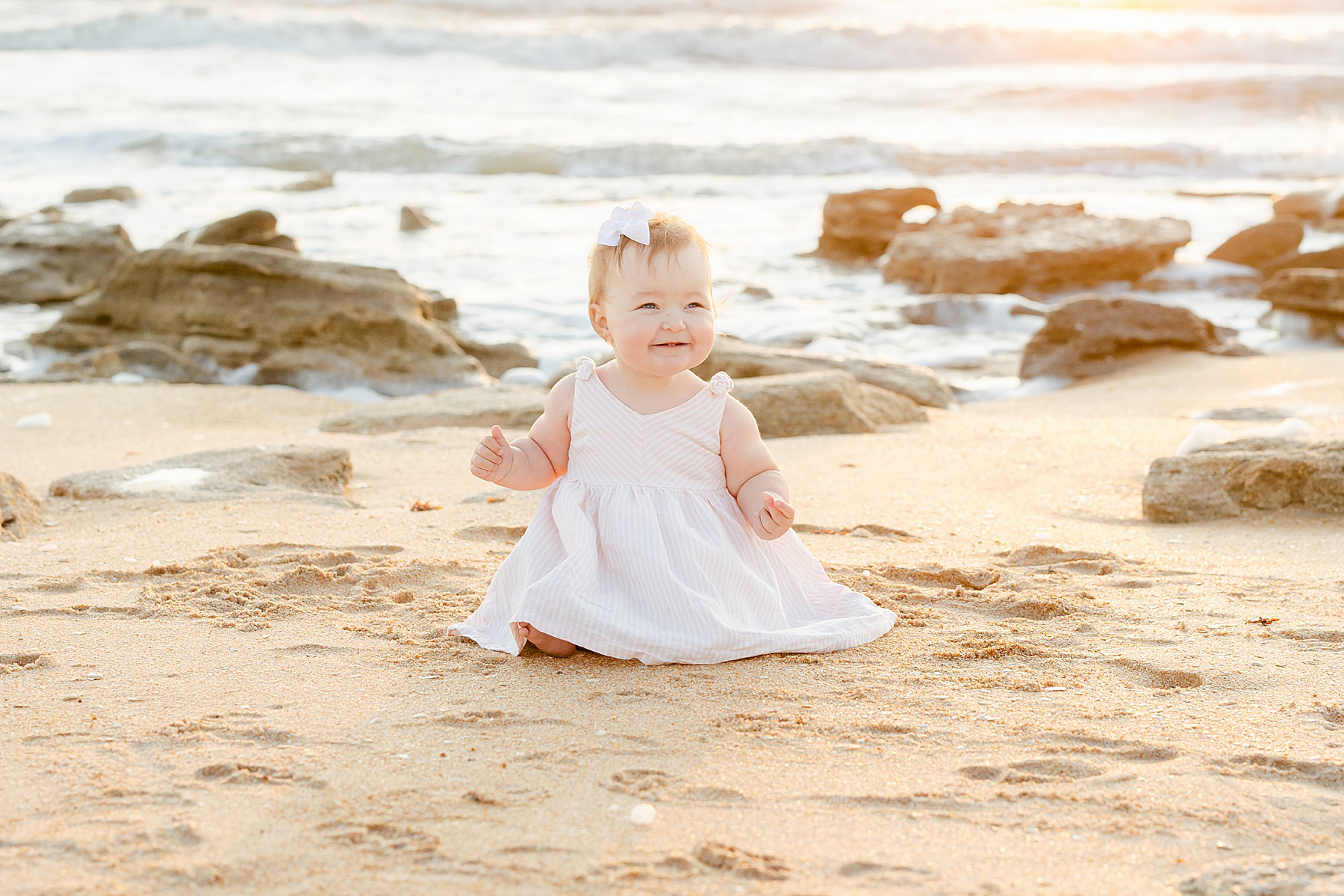 baby girl wearing pink white striped dress sitting on sand at sunrise beach