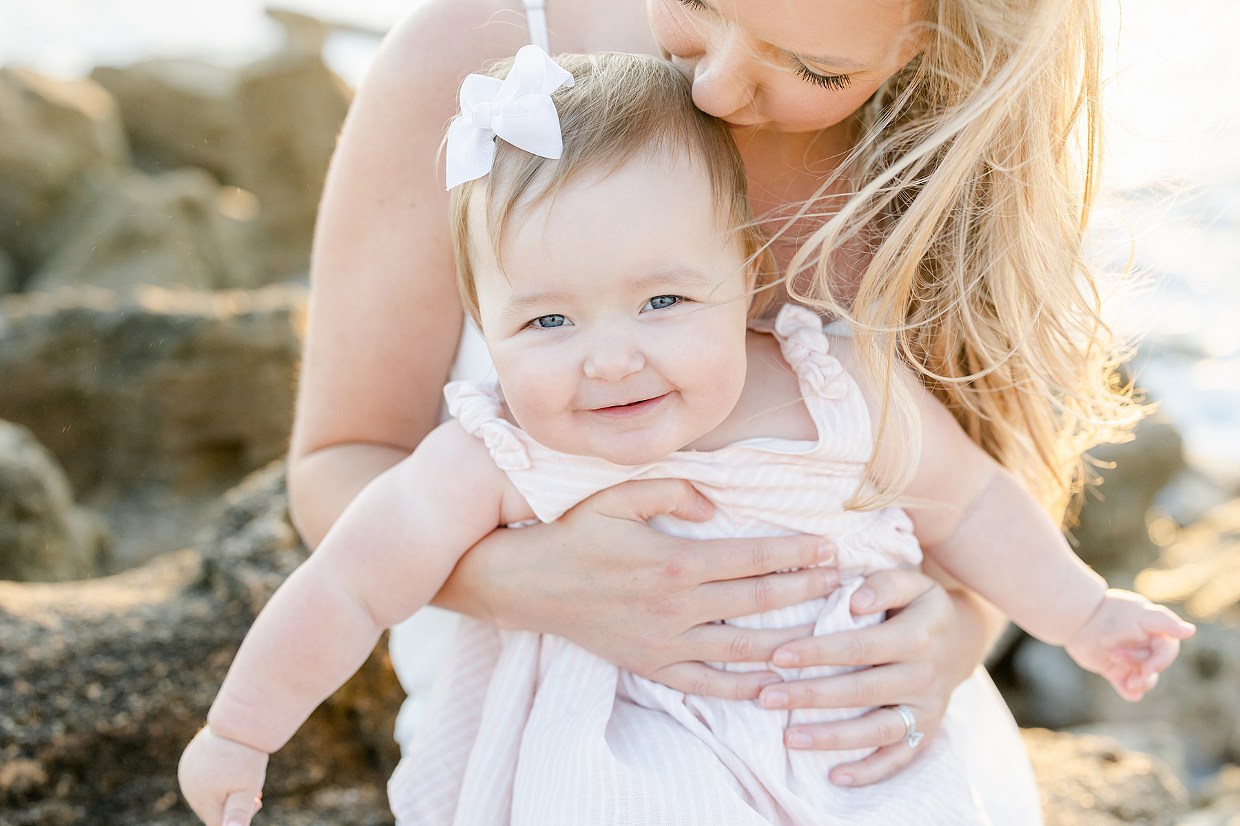 baby girl with white bow in hair wearing pink white striped dress on the beach at sunrise with mother with long blond hair