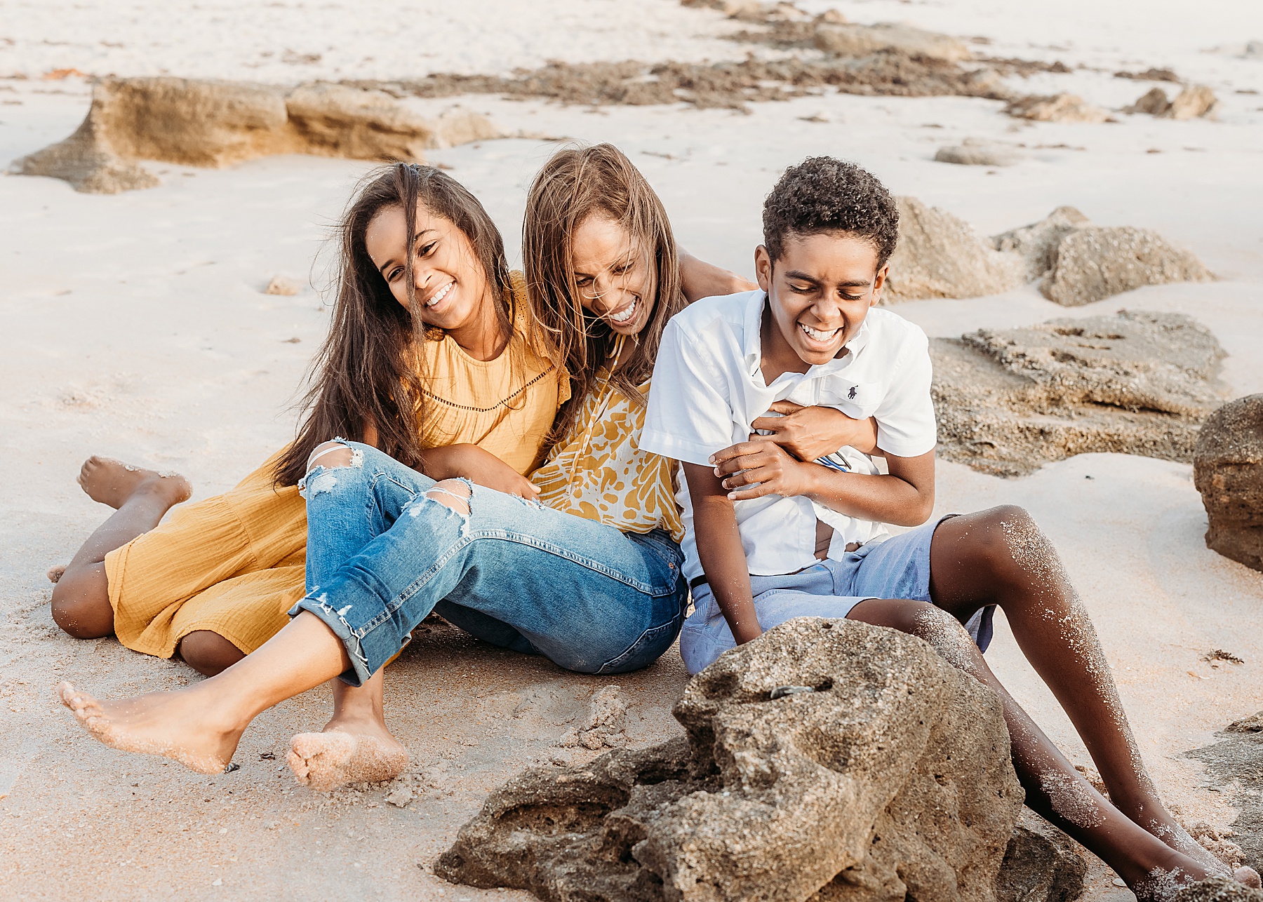 family of three sitting together on the sand at the beach near rocks