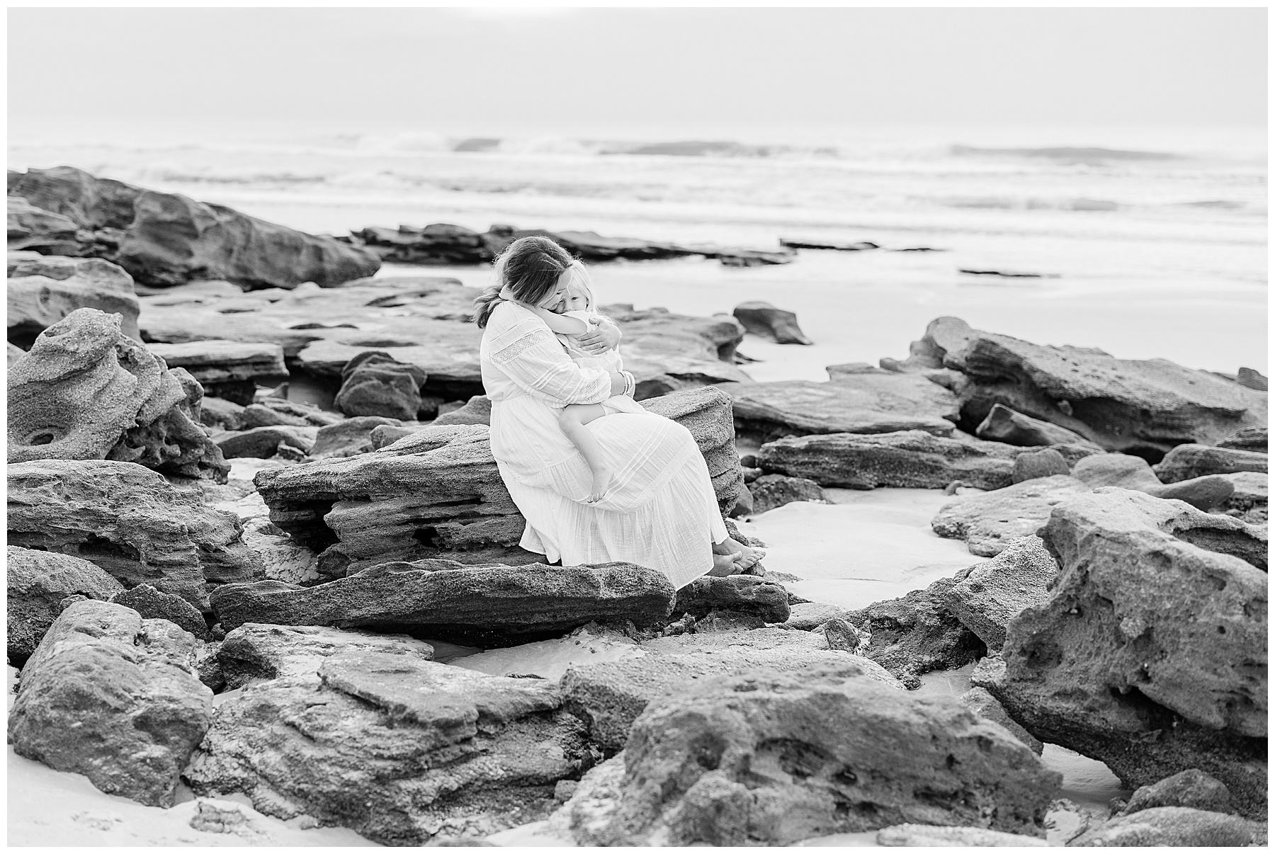 black and white image of mom holding little girl on the rocks on the beach