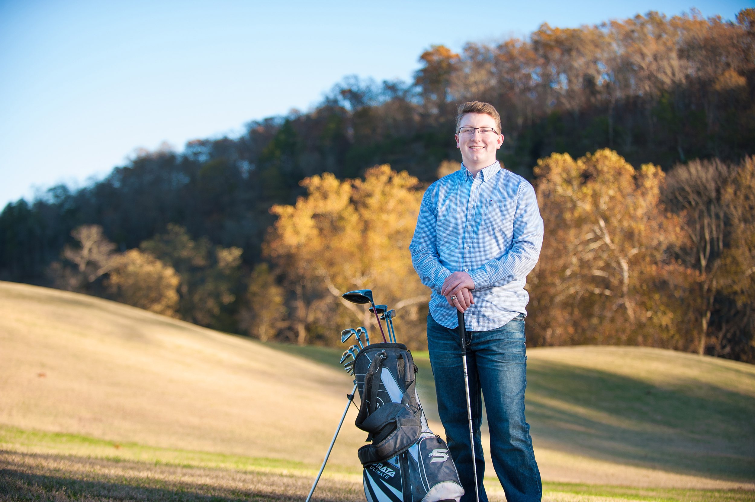 Senior guy in blue shirt and jeans standing on golf green with fall foliage behind him.