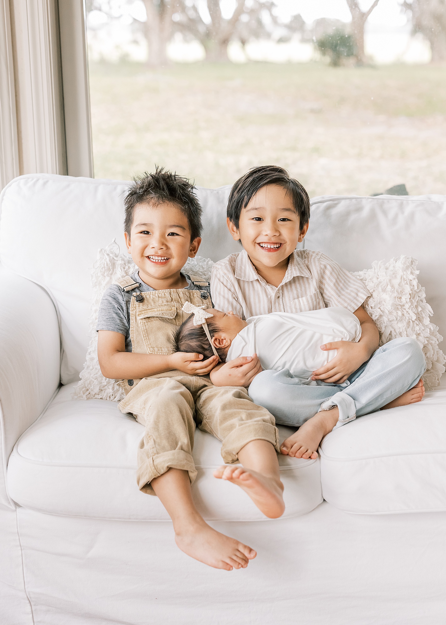 little boys sitting on white couch smiling while holding their newborn baby sister in Gainesville Florida