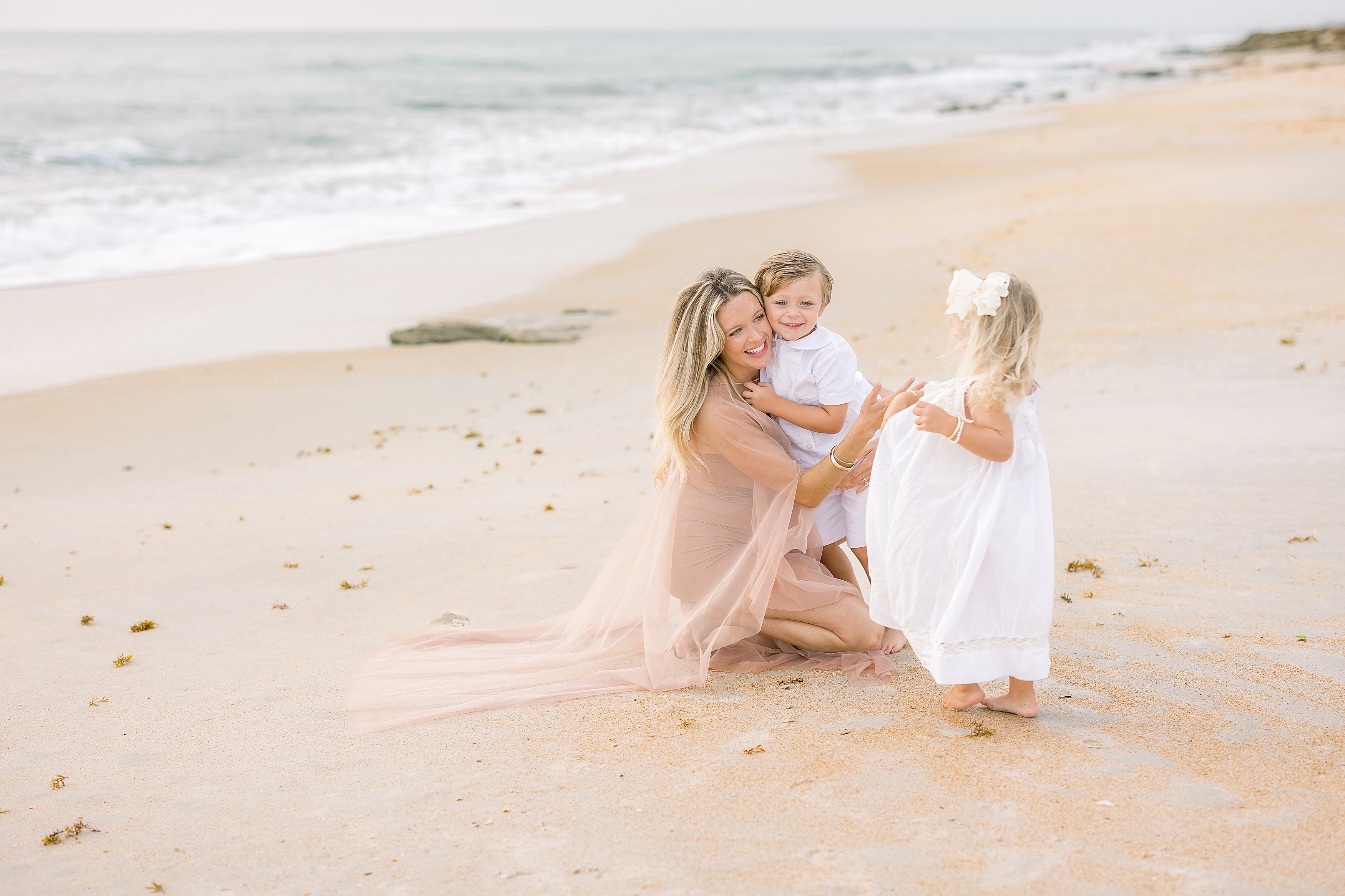 pregnant woman dressed in neutral clothing laughing with children on the beach at sunrise