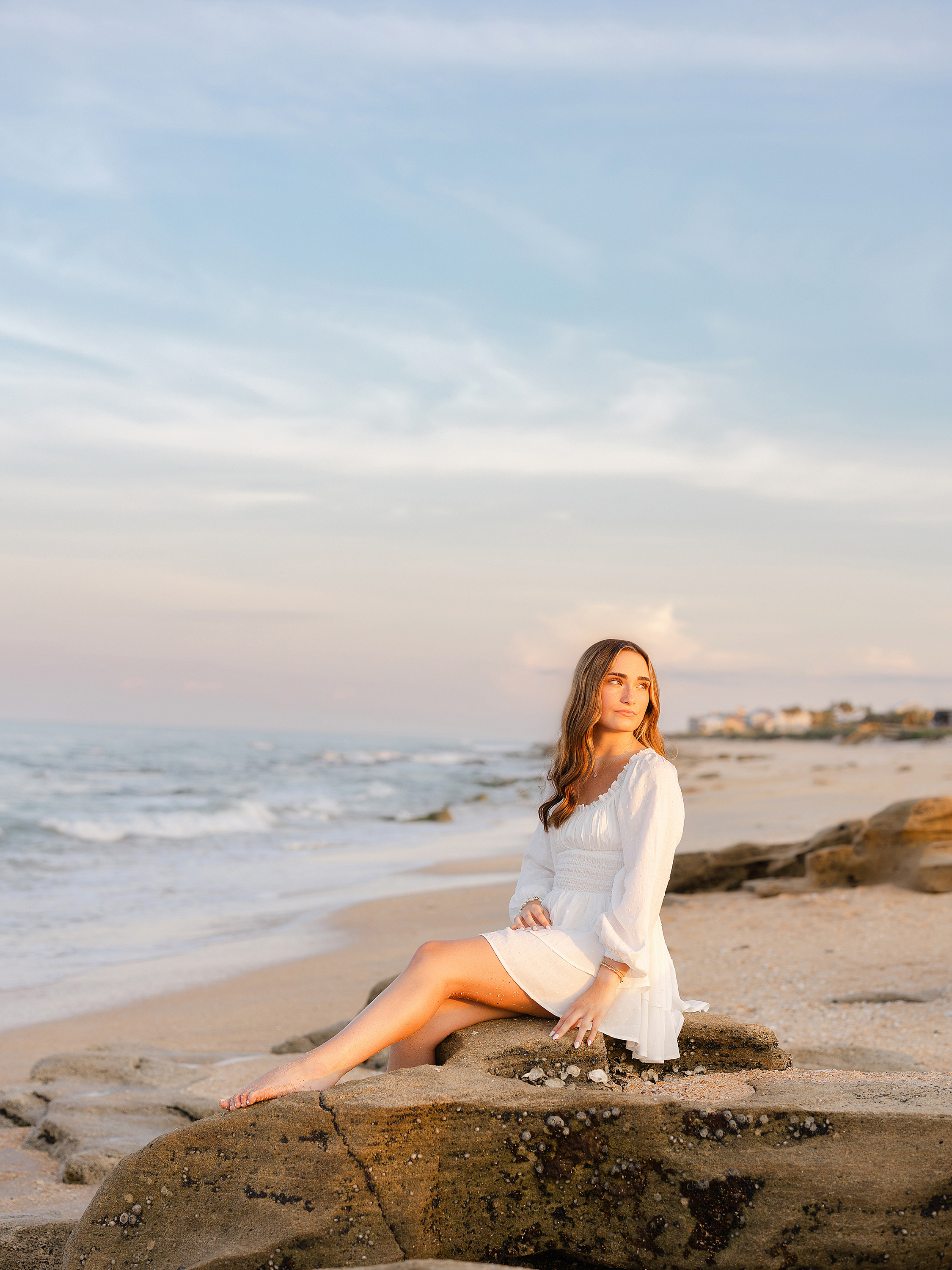Beach portrait of young woman in white dress near the shoreline during a pastel sunset.